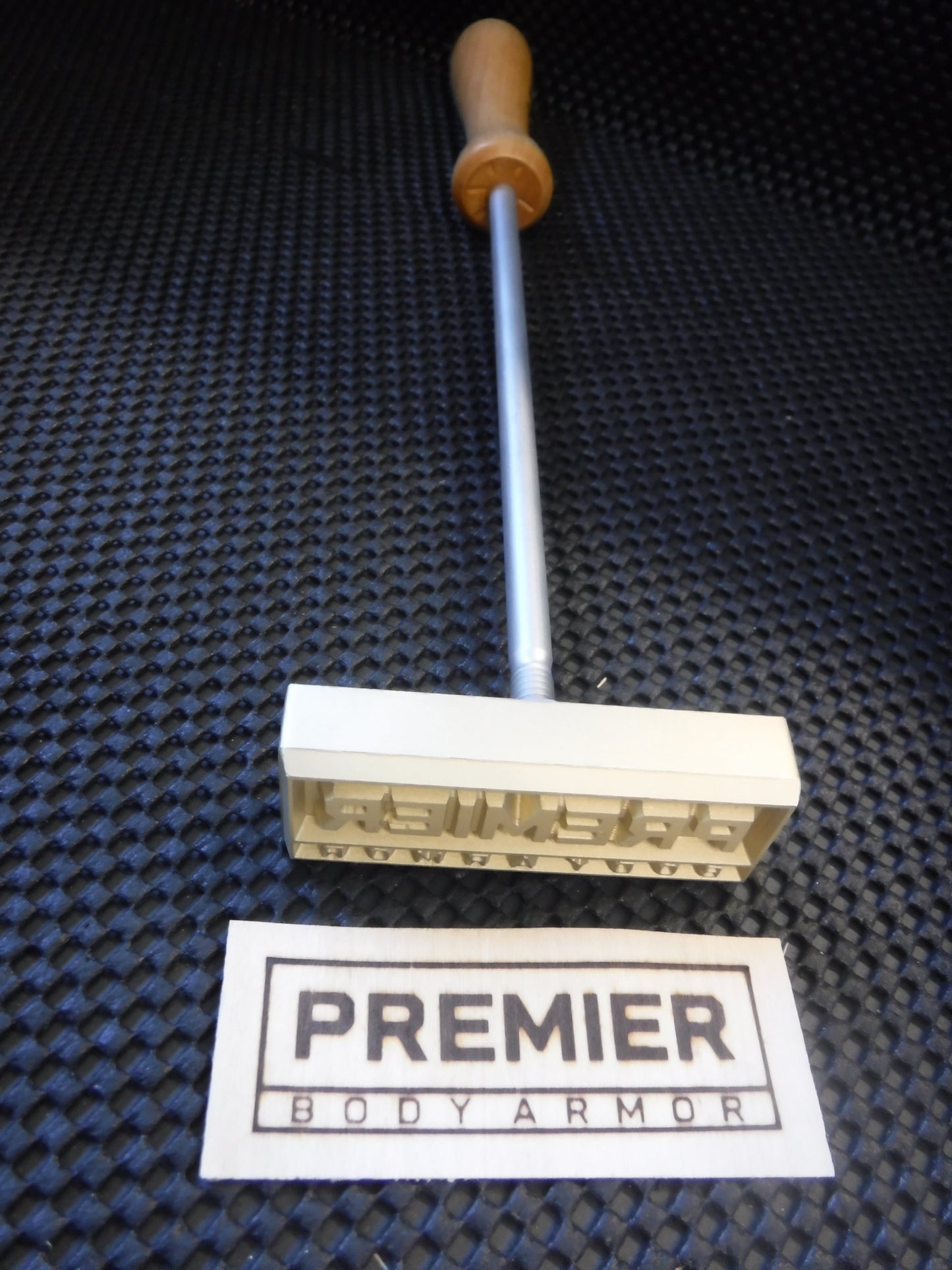 Metal stamp with wooden handle and 'Premier Body Armor' label on a textured black surface